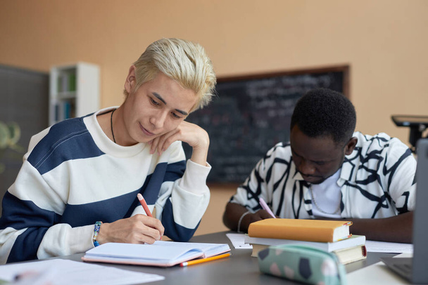 Молодой азиат с окрашенными светлыми волосами просматривает тестовые вопросы на бумаге, сидя за столом и записывая ответы в блокнот - Фото, изображение