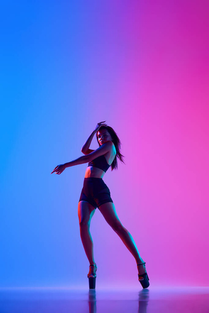 Piękna tancerka nosząca body i szpilki podczas wykonywania trików tanecznych na rurze w kolorowym świetle neonowym. Koncepcja stylu tańca, nowoczesny styl, współczesny, kultura młodzieżowa, autoekspresja - Zdjęcie, obraz