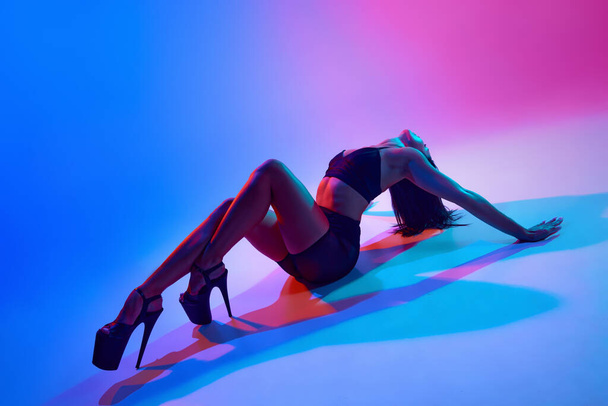 Молодая красивая стройная танцовщица на высоких каблуках позирует на полу в танцевальной студии в сине-розовом неоновом свете. Стиль современного танца, искусство, эстетика, хобби, творчество. - Фото, изображение