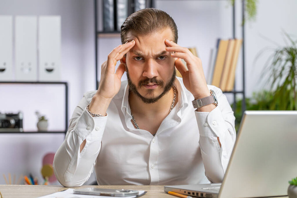 頭痛の問題の緊張片頭痛に苦しむ病気の中東のビジネスマンは,ホームオフィスデスクの職場でのストレス. ベアードフリーランサーブローカーの労働者はノートパソコンのノートPCで働く. オーバーワーキング - 写真・画像