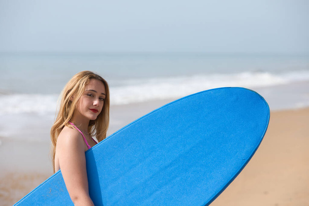 Nuori ja kaunis blondi surffaaja nainen vaaleanpunaisissa bikineissä ja sinisessä surffilaudassa. Tyttö nauttii lomastaan rannalla harjoittelemaan lempiurheilulajejaan. Matkailu ja loma käsite - Valokuva, kuva