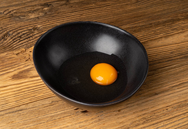 Jedno rozbite jajko w czarnej misce, surowe żółtko i białe, świeże rozbite organiczne jajko kurze na drewnianym rustykalnym tle - Zdjęcie, obraz