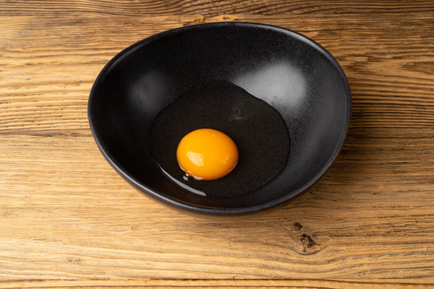 Un œuf cassé dans un bol noir, jaune cru et blanc, oeuf de poulet biologique frais cassé sur fond rustique en bois - Photo, image