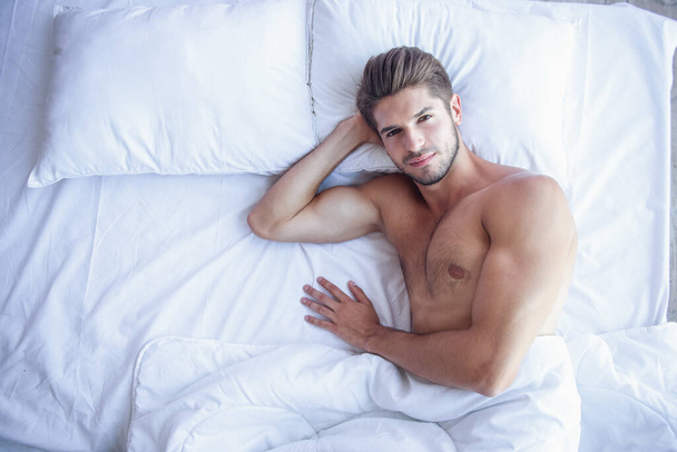 Bovenaanzicht van sexy gespierde jongeman die op zoek naar camera met sensuele glimlach terwijl liggend in bed - Foto, afbeelding