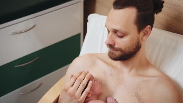 Padre sentado en un sillón en el hospital vinculándose con un hijo recién nacido. Vista desde arriba del nuevo padre americano cuidando a su bebé recién nacido haciendo de piel a piel en el hospital en el departamento postnatal - Metraje, vídeo