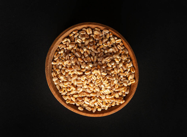 Пшеничные зерна в чаше из дерева, ячменная куча, сухие зерновые семена для хлеба, Спелта здоровой органической пищи, пшеничные зерна куча на черном фоне - Фото, изображение