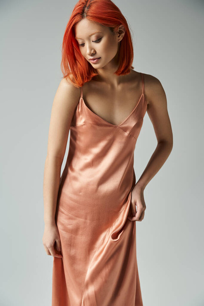 elegant young asian woman with red hair posing in silk and pastel pink slip dress on grey background - Photo, Image