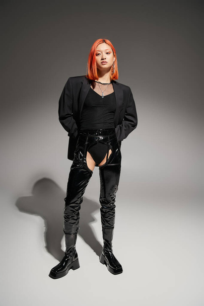 full length of asian woman in sexy latex boots and dark blazer standing on grey backdrop, bold style - 写真・画像