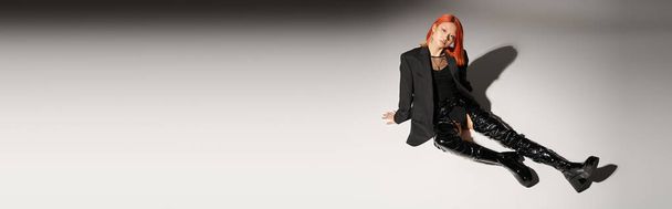 full length of sassy asian woman in sexy latex boots and blazer sitting on grey backdrop, banner - Photo, Image