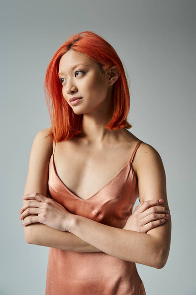 smiling young asian woman with red hair posing in pastel slip dress on grey backdrop, looking away - Foto, Imagen