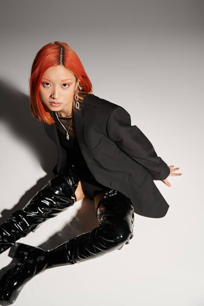 high angle of sassy asian woman in sexy latex boots and blazer sitting on grey backdrop, bold style - Fotografie, Obrázek