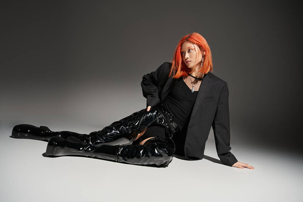 edgy style, asian woman with red hair sitting with hand on hip and latex boots on grey background - Photo, Image