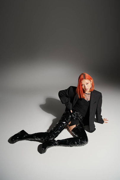 captivating asian woman with red hair sitting with hand on hip and latex boots on grey background - Photo, Image