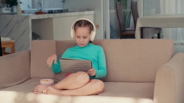 Medium long shot of pre-teen girl watching videos in social net on digital tablet while resting on sofa with snacks in studio flat - Footage, Video