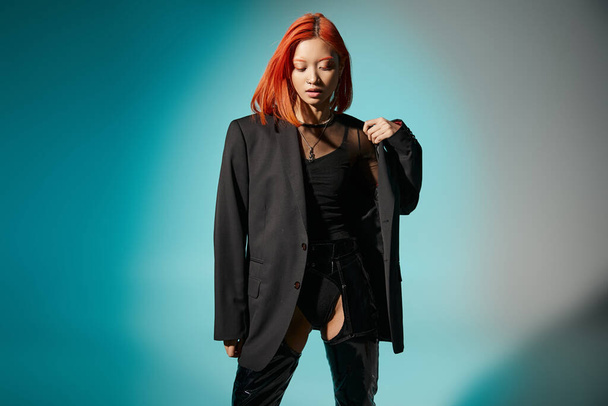 stylish asian model with red hair posing in blazer and black latex high boots on blue background - Photo, Image