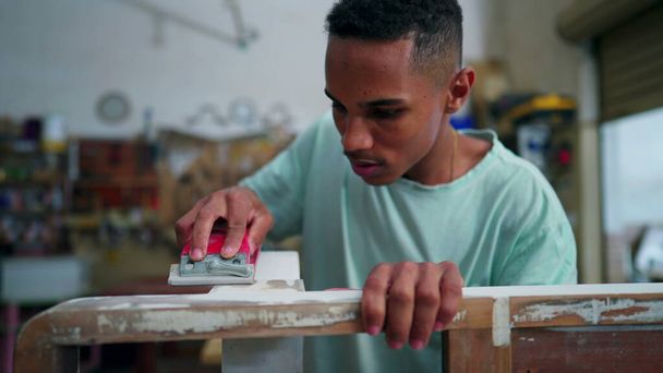 Um jovem carpinteiro brasileiro preto poliu a superfície de madeira na oficina de carpintaria com ferramenta. Empregado envolvido com a ocupação do trabalho, focado na reparação de móveis - Foto, Imagem
