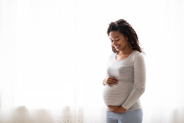 Těhotenská koncepce. Portrét usmívající se mladé černošky těhotné ženy objímající břicho při stání v blízkosti okna doma, afroameričanky Očekávající dáma něžně dotýkající se břicha, kopírovat prostor - Fotografie, Obrázek