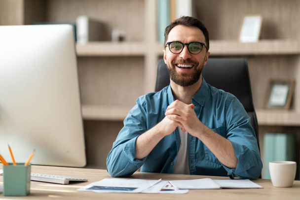 Modern üzletember. Portré jóképű fiatalember pózol az irodában, boldog Y-generációs férfi vállalkozó visel szemüveg ül a munkahelyen, nézi a kamera és mosolygós - Fotó, kép