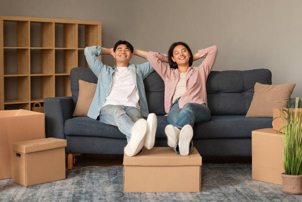 Neuer Eigenheimkauf. Entspannte asiatische Ehepartner, die sich auf dem Sofa zwischen gepackten Kisten ausruhen und den beweglichen Tag im Haus genießen, sitzen mit geschlossenen Augen und halten die Hände hinter dem Kopf. Konzept zur Vermietung von Immobilien - Foto, Bild