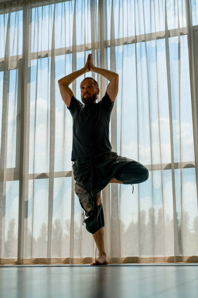 yogui hombre de pie en una pierna con las manos arriba y realiza ejercicios de yoga kundalini en una hermosa sala de ejercicios - Foto, Imagen