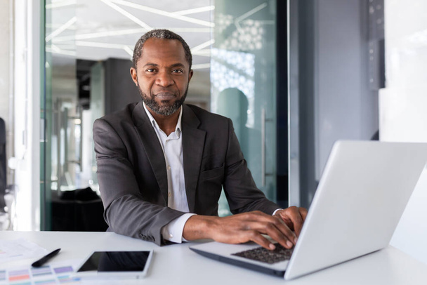 Portrait de pensée sérieuse et homme d'affaires confiant, patron afro-américain pensant et regardant la caméra, homme travaillant à l'intérieur du bureau en costume d'affaires, investisseur financier avec ordinateur portable. - Photo, image