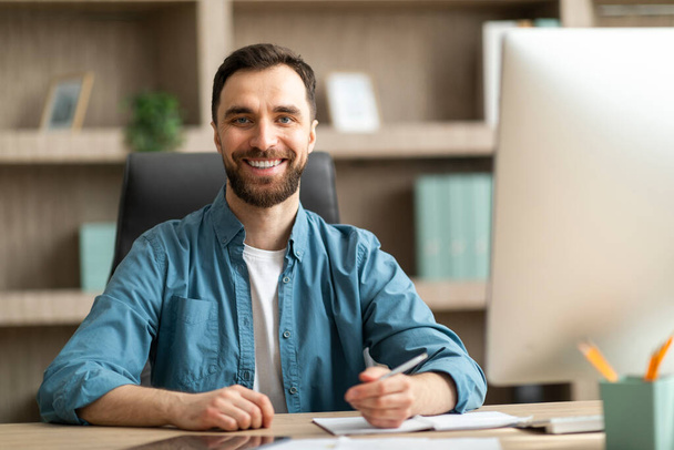 Osoba biznesowa. Portret przystojnego młodego biznesmena pozującego w biurze, millenijny przedsiębiorca siedzący przy biurku i robiący notatki, patrzący i uśmiechający się do kamery, kopiujący przestrzeń - Zdjęcie, obraz