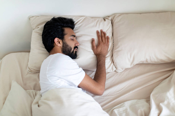 Rauhallinen nuori intialainen mies nukkuu mukavalla sängyllä kotona, komea tuhatvuotinen itämainen kaveri torkkuu pehmeällä tyynyllä peiton alla makuuhuoneen sisustuksessa, nauttien terveellisestä unesta, ylhäältä katsottuna vapaalla tilalla - Valokuva, kuva
