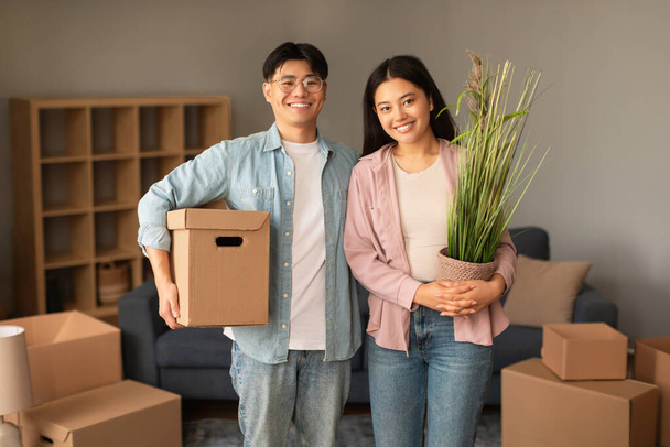 Perheen muuttotalo. Iloinen kiinalainen nuori pari poseeraa pakattujen pahvilaatikoiden ja kasvien kanssa potissa, valmiina siirtymään ja siirtymään uuteen kotiin hymyillen kameralle. Oma asunto Kiinteistön osto - Valokuva, kuva