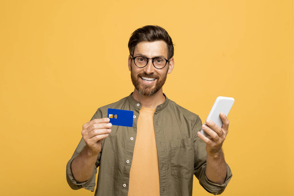 Virtueel bankieren, e-commerce. Gelukkig europees man van middelbare leeftijd met smartphone en bank credit card in zijn handen poseren op gele achtergrond, glimlachen op camera - Foto, afbeelding