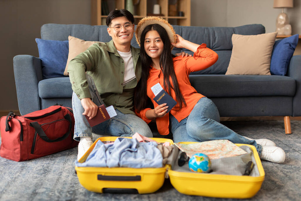 Urlaubsreisen. Fröhliches junges koreanisches Paar mit Fahrkarten und Reisepässen in der Nähe geöffneter Koffer im Innenraum, die Urlaubsplanung lächelnd in die Kamera. Schuss von Weltenbummlern Ehepartner bereit für neue Reise - Foto, Bild