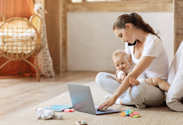 Geschäftsfrau mit Baby, die von zu Hause aus mit Laptop und Handy arbeitet. Junge Mutter sitzt mit Kleinkind im Arm auf dem Boden, telefoniert und tippt am PC, kopiert Leerzeichen - Foto, Bild