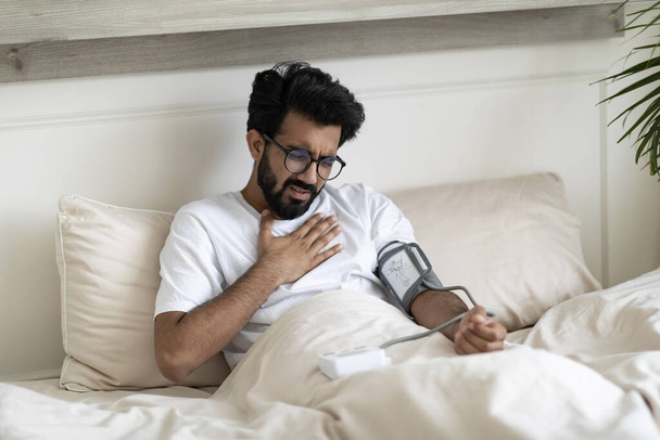 Health Problems. Young Indian Guy Having Chest Pain At Home, Checking Blood Pressure With Upper Arm Monitor While Sitting In Bed At Home, Sick Eastern Man Measuring Arterial Tension, Copy Space - Фото, зображення