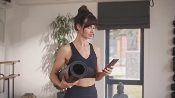 Beautiful Caucasian female fitness trainer chatting with her customer on phone while standing in sport center. Sporty woman holding mat during remote communication. Gym concept. - Footage, Video