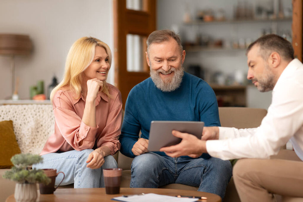 Agent Man Showing Digital Tablet To Excited Mature Clients Couple Offering His Digital Services, Sitting On Sofa In Their Living Room Indoor. Real Estate Buyers Choosing House Offers With Dealer - Photo, Image