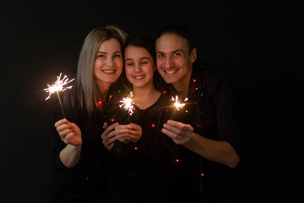 Happy family in black - woman, man and little girl, with a sparkler in hand the New Years Christmas at home. The girl at the father on hands. New year 2023 - Fotografie, Obrázek