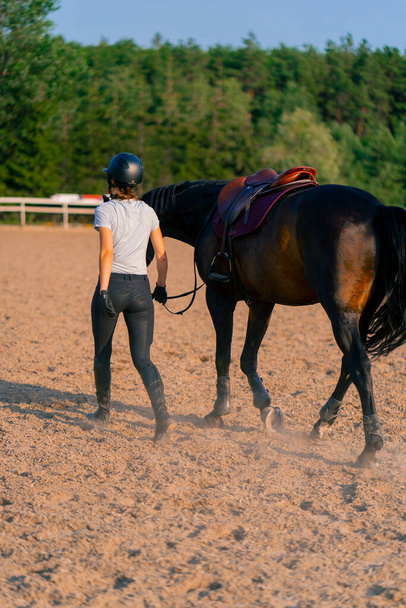 Шоломована гонщик веде її красивий чорний кінь біля в'язання на верховій арені під час верхової їзди - Фото, зображення