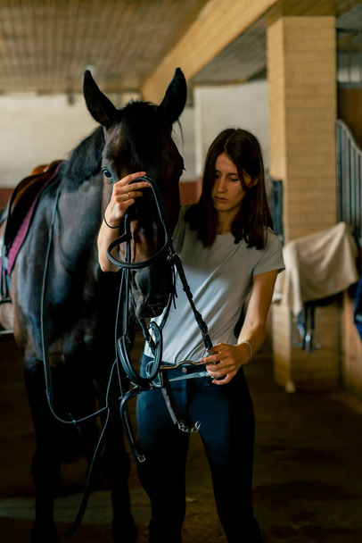 Un cavaliere mette l'imbracatura sul suo cavallo nero nelle scuderie in preparazione al concetto di corsa equestre. - Foto, immagini