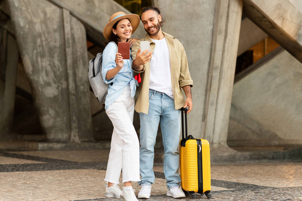 Cheerful young european man with beard, lady hold passport, take selfie on smartphone, enjoy trip on station. Tourist emotions, active lifestyle, summer vacation, photo for blog and social networks - Photo, Image