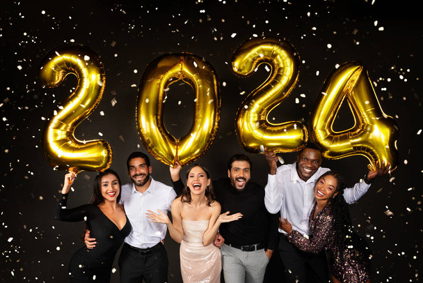 Щасливі багаторасові чоловіки і жінки три люблячі тисячолітні пари одягнені в гарні вбрання, що святкують новий 2024 рік разом на чорному тлі, тримаючи золоті числові кульки і посміхаючись на камеру - Фото, зображення
