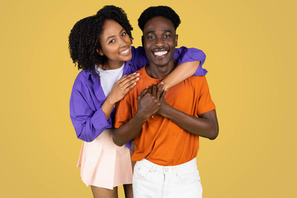 Cheerful millennial black lady hugging boyfriend in casual, enjoy success, date, isolated on yellow studio background. Human emotions, lifestyle, ad and offer, love and relationships - Photo, Image