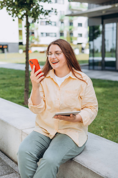 Schöne junge Frau, die draußen auf der Straße sitzt und ihr Handy benutzt. Urban Lifestylekonzept. Reisende, Online-Bildung, Geschäft oder Einkaufen - Foto, Bild