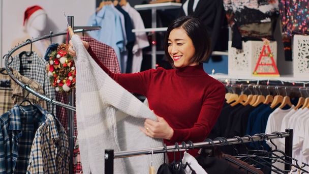 Asiatische Kunden stöbern durch Kleiderständer im weihnachtlich dekorierten Geschäft und prüfen die passende Kleidungsgröße. Frau in Weihnachtskleidung schmückt Modeboutique im Winter - Foto, Bild
