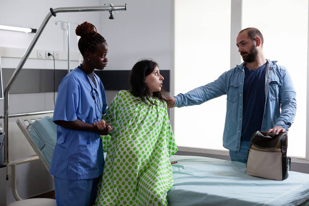 Afro-Amerikan doğum hemşiresi hamile bir kadının hastane koğuşunda yataktan kalkmasına yardım ediyor, hastayı sezaryen ameliyatına hazırlıyor. Gelecekteki ebeveynler doğum kliniğinde çocuk bekliyor. - Fotoğraf, Görsel