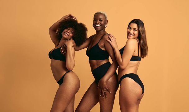 Side view of happy three multiracial females wearing black underwear and showing body positivity in studio - Foto, Imagem