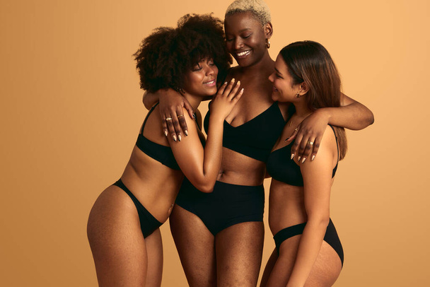 Group of multiethnic women in black lingerie embracing with eyes clothes while standing against beige background - Фото, зображення