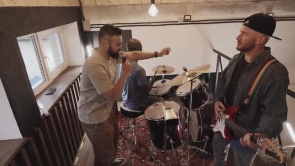 High angle medium full shot of three multiracial friends, black man playing on drums, smiling, Caucasian man in black cap playing guitar, another Caucasian man singing into microphone. Slow motion - Footage, Video