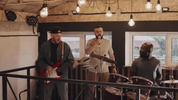 Medium full shot of three multiracial friends playing in band, black man playing on drums, Caucasian man in black cap playing guitar, both smiling, another Caucasian man singing into mic. Front view - Footage, Video