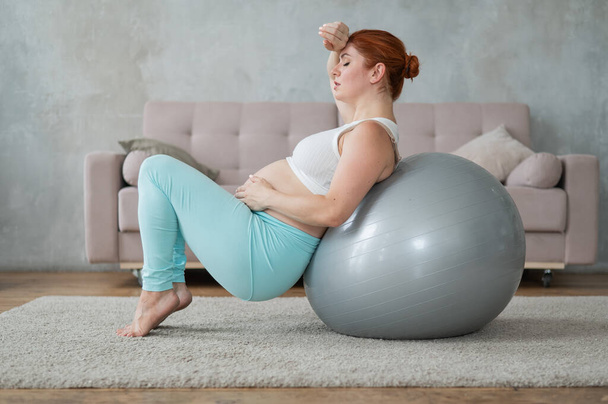 A pregnant woman lies with her back on a fitball and keeps her hands on her tummy at home. Expectant mother is in pain - Φωτογραφία, εικόνα