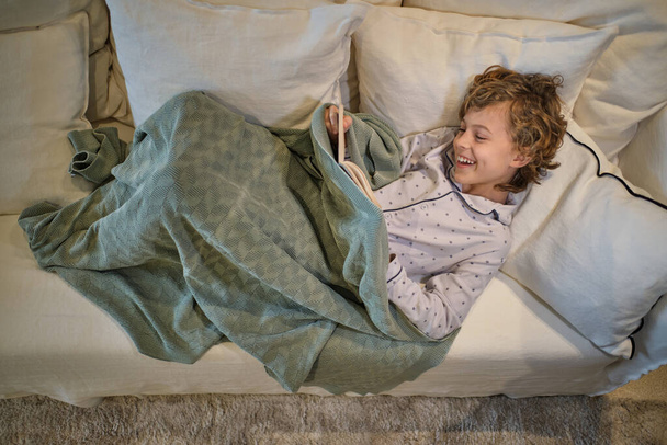 Od shora plné tělo veselého chlapce v pyžamu čtení knihy, zatímco odpočívá na pohodlném gauči v lehkém obývacím pokoji - Fotografie, Obrázek