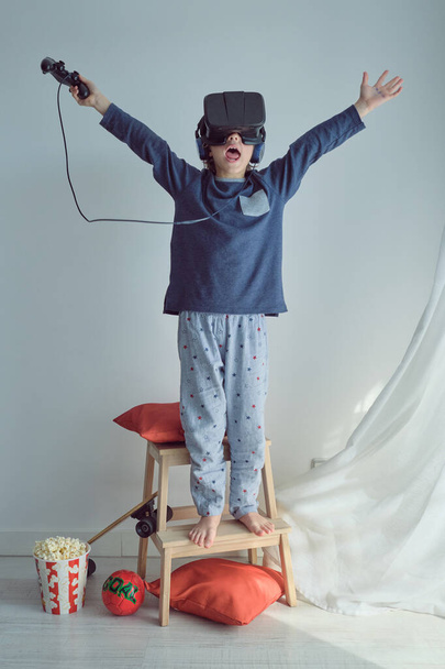 Pleine longueur de gamin anonyme joyeux avec la bouche ouverte levant les bras tout en se réjouissant du succès dans le jeu de réalité virtuelle portant casque et en utilisant contrôleur et écouteurs sur fond blanc - Photo, image